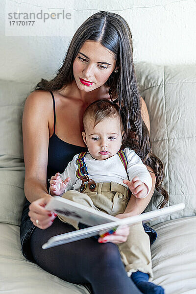 Mother reading book while sitting with baby boy on bed at home