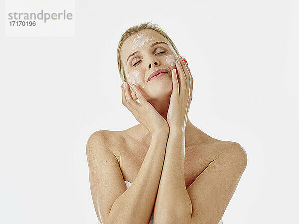 Woman wrapped in towel applying face cream while standing against white background