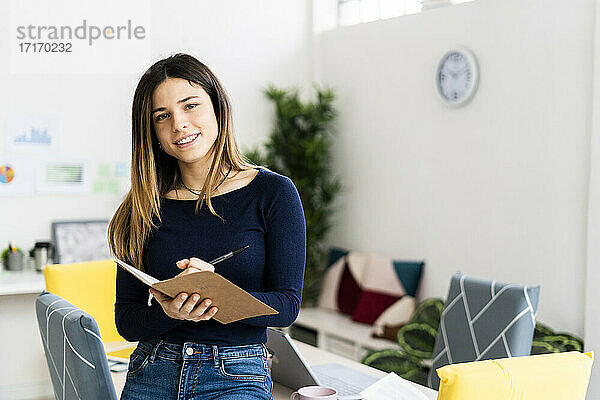 Lächelnde Studentin  die ein Notizbuch und einen Stift hält  während sie im Wohnzimmer steht