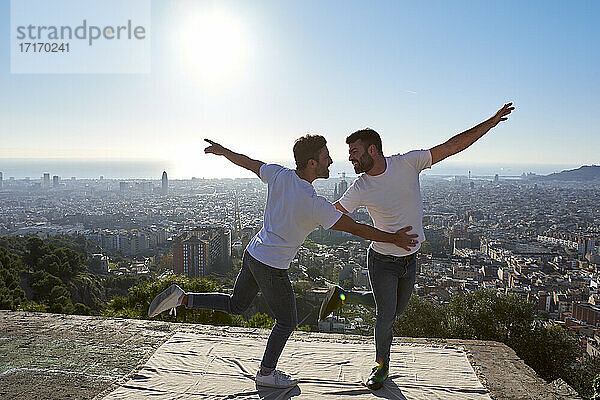 Glückliches schwules Paar mit ausgestreckten Armen tanzt auf dem Aussichtspunkt gegen den klaren Himmel  Bunkers del Carmel  Barcelona  Spanien