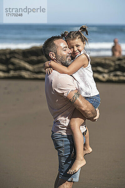 Smiling daughter embracing father at beach during sunny day