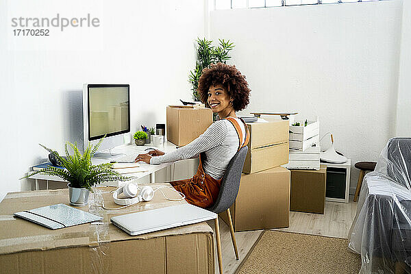 Lächelnde Frau sitzt auf einem Stuhl neben einem Computer  während sie in eine neue Wohnung umzieht