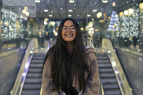 Cheerful young woman standing against illuminated glass wall in shopping mall