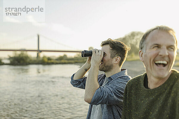 Son looking through binoculars while father laughing by at riverbank