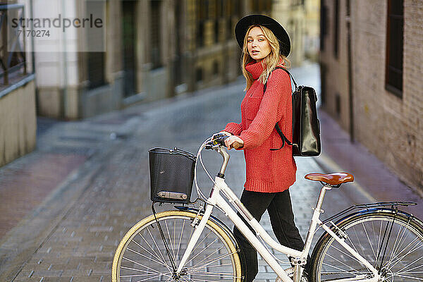 Woman wearing hat looking away while walking with bicycle on footpath in city