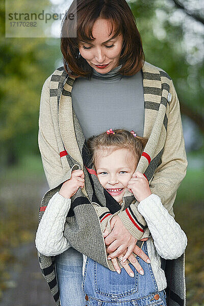 Glückliches Mädchen versteckt sich in Mutters Strickjacke im Park