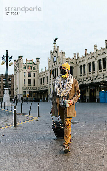 Modischer Mann mit Gesichtsmaske hält Kaffeetasse beim Spaziergang in der Stadt