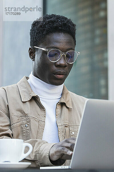 Young man in eyeglasses using laptop at sidewalk cafe