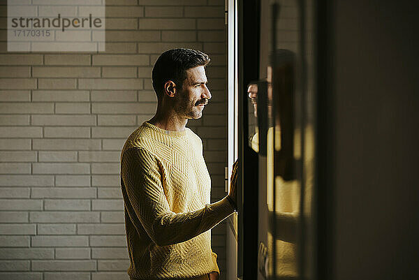 Man in yellow sweater looking through window at home