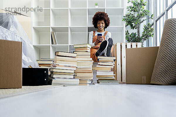 Afro-Frau  die ihr Smartphone benutzt  während sie in ihrem neuen Zuhause am Bücherregal sitzt