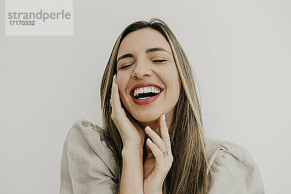 Laughing woman touching cheek with eyes closed against white background