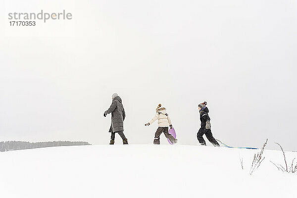 Vater und Kinder gehen mit Schlitten auf schneebedecktem Land gegen den Himmel