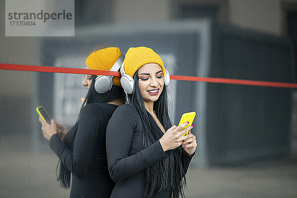 Beautiful woman with headphones over knit hat using smart phone against glass