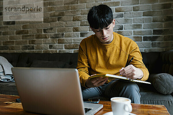 Junger Mann mit Laptop  der in ein Buch schreibt  während er auf dem Sofa im Café sitzt
