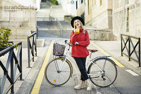 Young woman wearing hat drinking juice while standing with bicycle on road in city