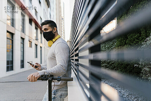 Young man with protective face mask standing by electric push scooter against metal fence while looking away during pandemic