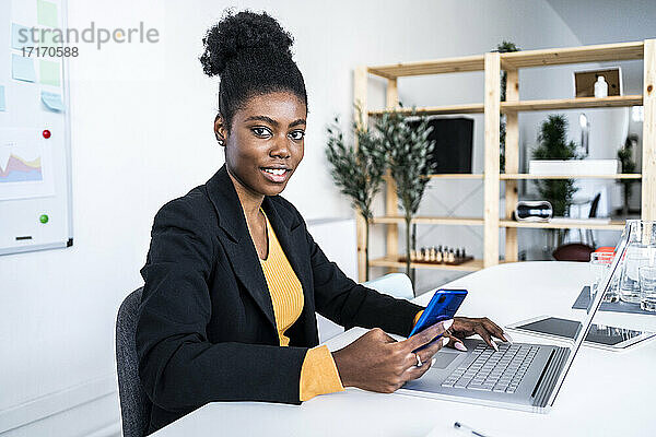 Young female Afro entrepreneur sitting with laptop and smart phone at desk in office