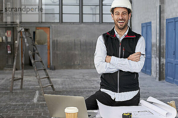 Männlicher Architekt mit verschränkten Armen  der lachend an einem Tisch in einem Gebäude steht