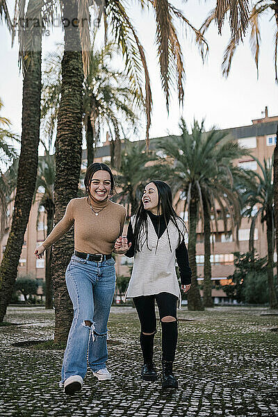 Fröhliche Freundinnen halten sich an den Händen  während sie auf einem Fußweg im Park laufen
