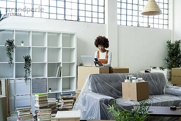 Afro-Frau  die beim Umzug in eine neue Wohnung ein Mobiltelefon mit Laptop benutzt