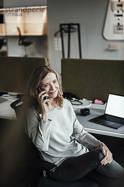 Lächelnde Geschäftsfrau im Gespräch über Smartphone bei der Arbeit am Schreibtisch im Büro