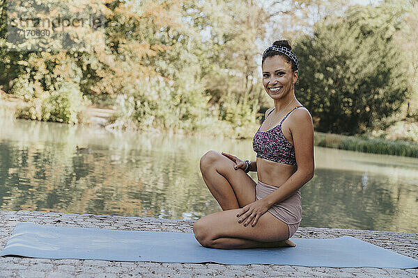 Female athlete smiling while sitting on exercise mat by lake at park