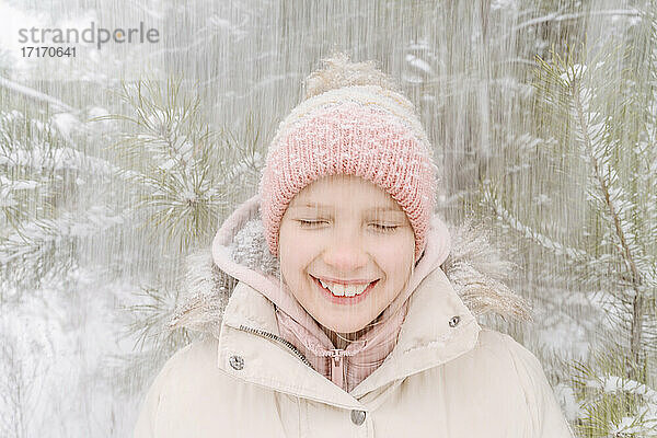 Nahaufnahme eines lächelnden Mädchens mit geschlossenen Augen  das den Schneefall im Wald genießt