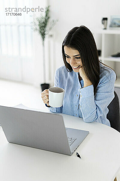 Berufstätige Frau trinkt Kaffee  während sie im Büro sitzt und einen Laptop benutzt