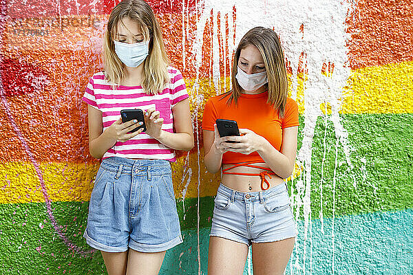 Modische Freundinnen in kurzen Hosen mit Smartphones an einer Graffiti-Wand während einer Pandemie