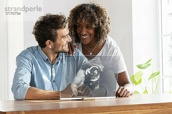 Lächelndes Paar bei der Überprüfung eines Ultraschallbildes auf einem transparenten Bildschirm zu Hause