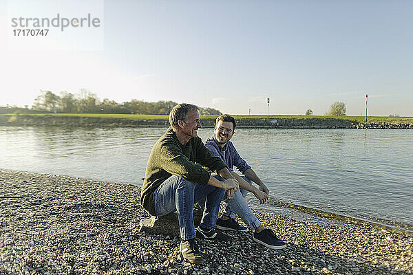 Father and son spending leisure time sitting at riverbank against clear sky
