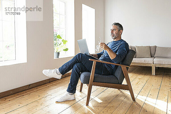 Nachdenklicher Geschäftsmann  der eine Brille hält  während er zu Hause am Laptop sitzt