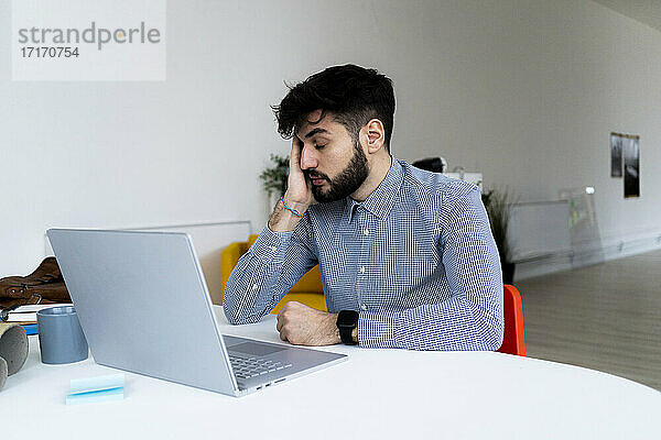 Müder Geschäftsmann mit Blick auf den Laptop im Kreativbüro