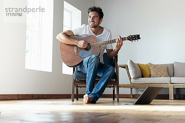 Lächelnder Mann spielt Gitarre  während er auf einem Stuhl zu Hause sitzt
