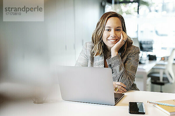 Lächelnde Geschäftsfrau mit Laptop auf dem Schreibtisch sitzend im Büro