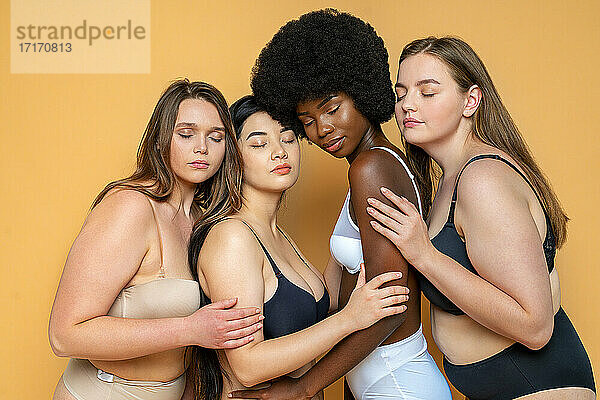 Multi-ethnische Gruppe von weiblichen Modellen in Dessous mit geschlossenen Augen  die sich vor gelbem Hintergrund umarmen
