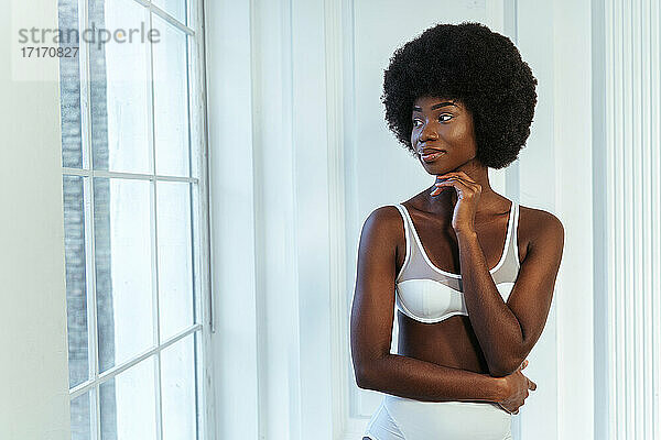Afro weibliches Modell in Dessous mit Hand am Kinn schaut durch Fenster gegen weiße Wand