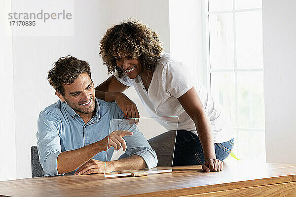 Lächelndes Paar berührt den Bildschirm eines transparenten Geräts  während es zu Hause sitzt