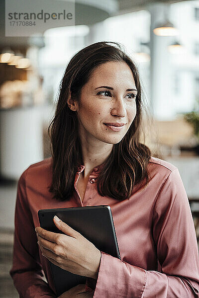 Lächelnde weibliche Fachkraft schaut weg  während sie ein digitales Tablet in einem Café hält