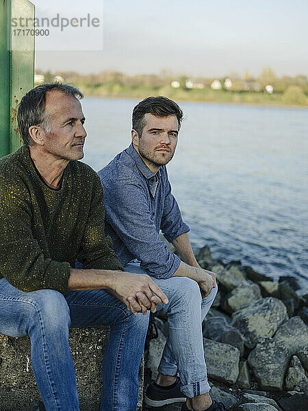 Father and son contemplating while sitting on rock against river
