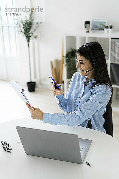 Geschäftsfrau mit Headset  die einen Notizblock mit ihrem Mobiltelefon fotografiert  während sie am Schreibtisch im Büro sitzt
