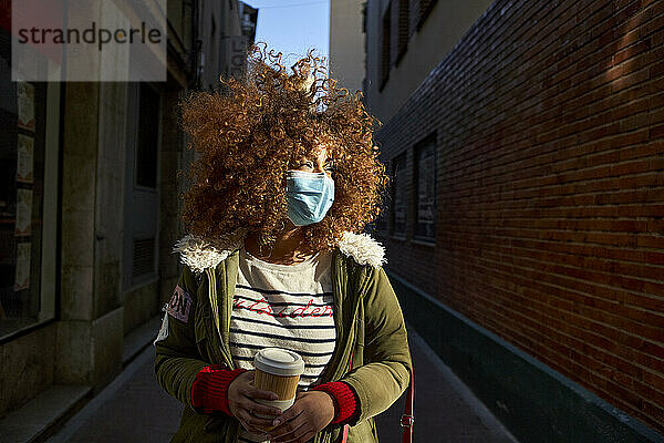 Brown curly hair woman wearing protective face mask looking away while standing with coffee cup on footpath