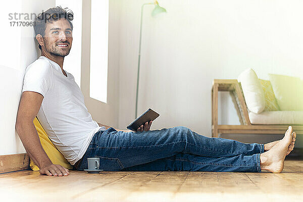 Junger Mann mit digitalem Tablet  der lächelnd zu Hause auf dem Boden sitzt