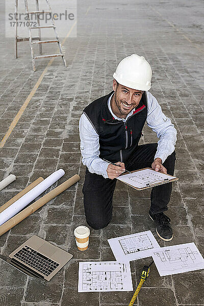 Lächelnder männlicher Architekt  der einen Lageplan hält  während er in einem Gebäude auf dem Boden kniet