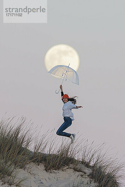 Joyful young woman with umbrella jumping on top of sand dune against sky during dusk