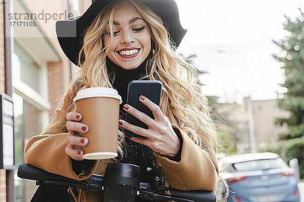 Happy young woman using smart phone while holding reusable coffee cup