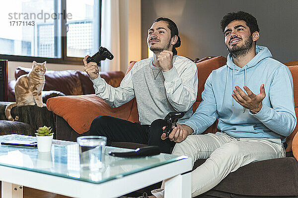 Junge männliche Freunde spielen ein Videospiel  während sie zu Hause auf dem Sofa im Wohnzimmer sitzen