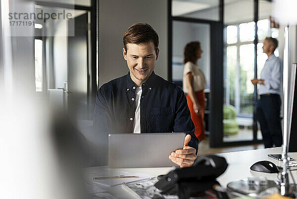 Lächelnder Geschäftsmann  der ein digitales Tablet benutzt  während er mit Kollegen im Hintergrund in einem Großraumbüro sitzt