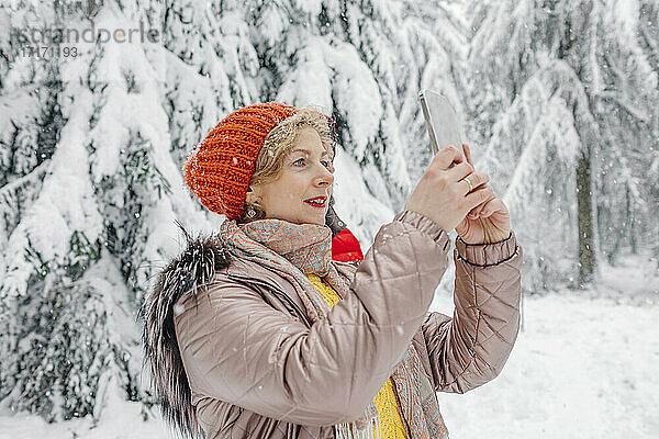 Ältere Frau in warmer Kleidung  die im Wald stehend ein Mobiltelefon benutzt