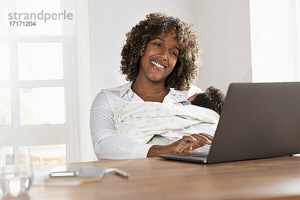 Lächelnde Mutter  die ihr Baby hält  während sie im Büro zu Hause am Laptop arbeitet